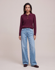 A woman stands against a plain beige studio background wearing a LeDoré Camille cable knit polo sweater with gold buttons, light blue wide-leg jeans, and dark brown shoes.