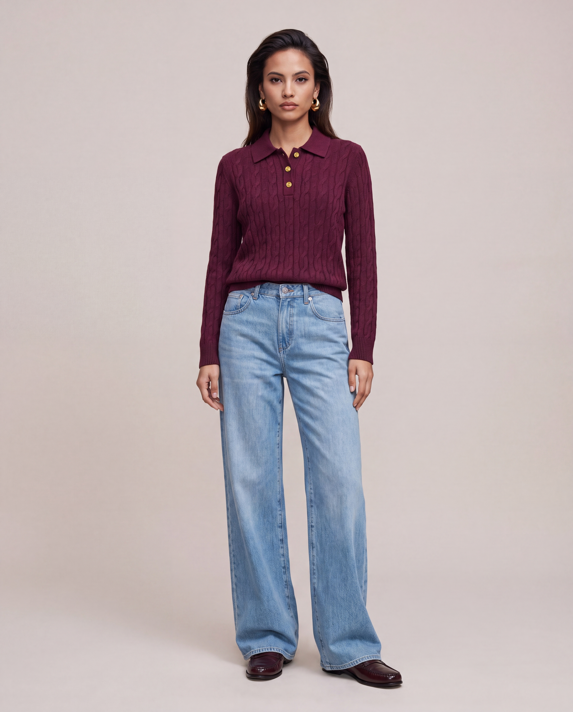 A woman stands against a plain beige studio background wearing a LeDoré Camille cable knit polo sweater with gold buttons, light blue wide-leg jeans, and dark brown shoes.